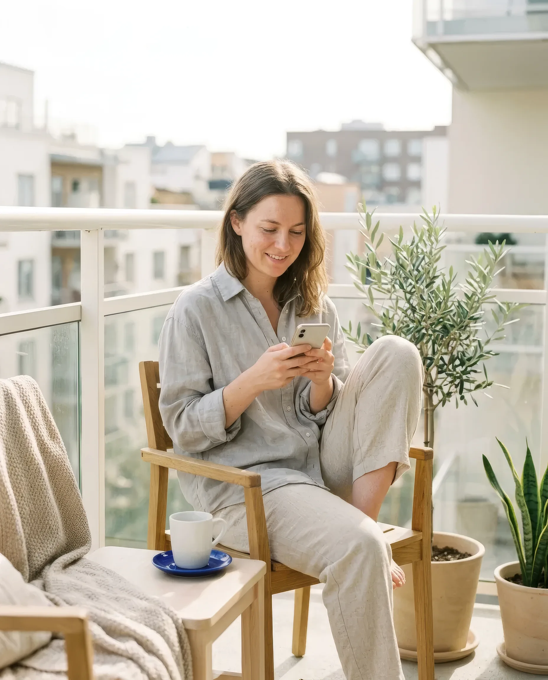 A woman in her late twenties on a sunny apartment balcony reading a study invitation on her phone, coffee beside her, smiling, candid morning light.