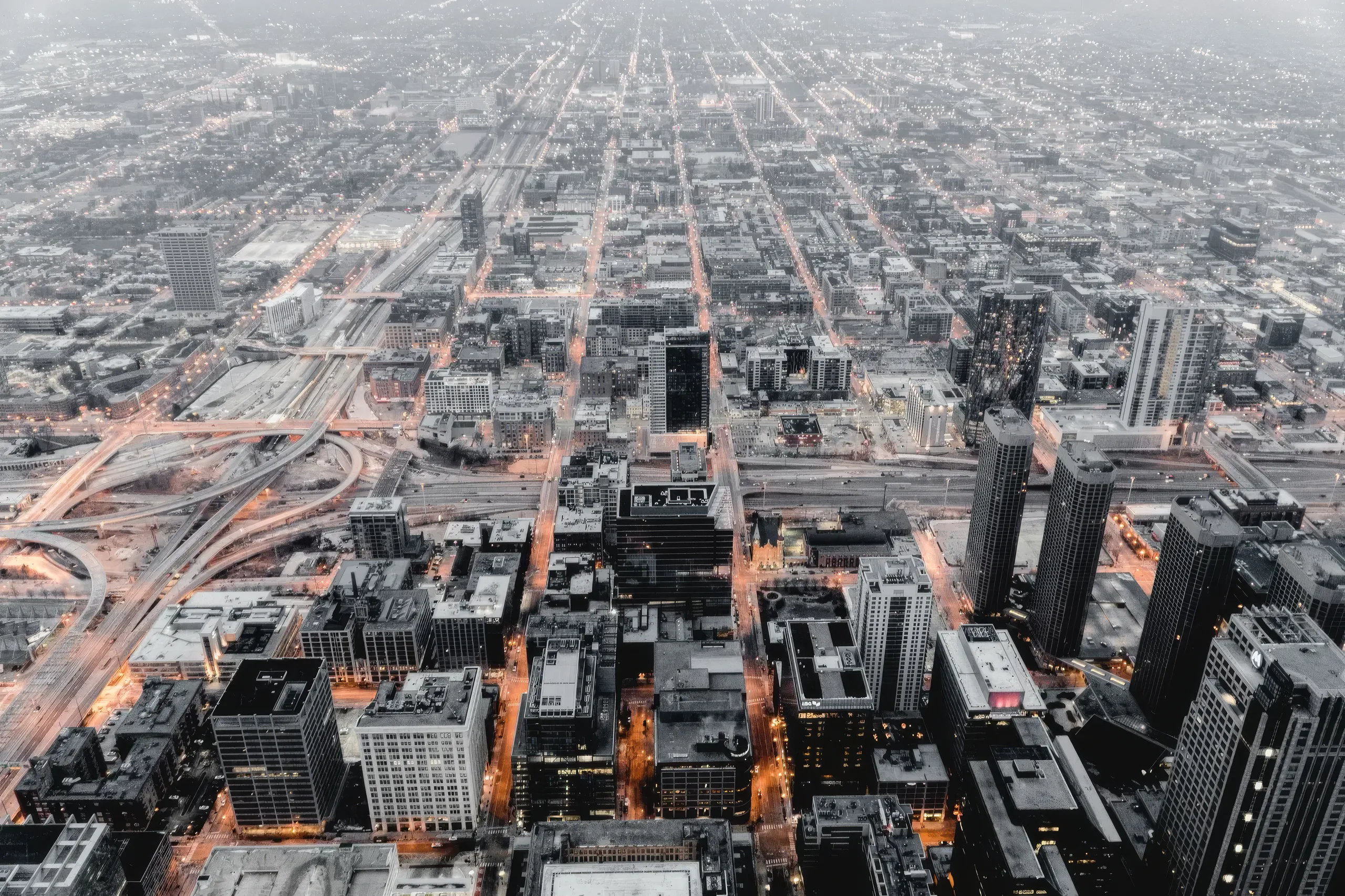 A city seen from high above, its grid of streets running out to a grey horizon, lit amber at dusk.